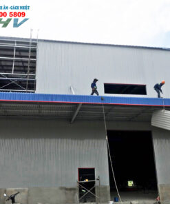 Xưởng xây dựng với hai công nhân đang thi công mái tôn màu xanh trên tòa nhà công nghiệp.