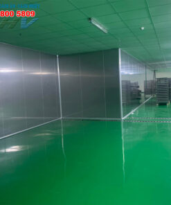 Phòng có sàn epoxy xanh, tường ốp tôn nhôm và kệ sắt trong nhà xưởng.